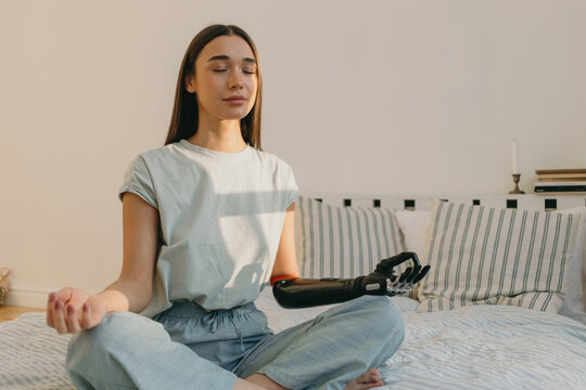 Pretty cyber lady with 3d-printed bionic hand prosthesis doing yoga sitting in lotus posture, fingers in mudra sign, having eyes closed, meditating on bed in morning, reaching zen and harmony