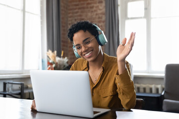 Cheerful happy African American hipster girl in headphones, glasses waving hand hello at webcam of...