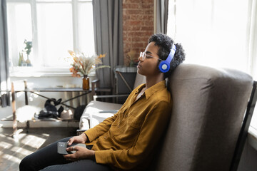 Obraz premium Peaceful calm millennial Afro American woman taking pause, enjoying lounge relaxing music on comfortable sofa at home, meditating with closed eyes, concentrating on song, sounds, holding smartphone