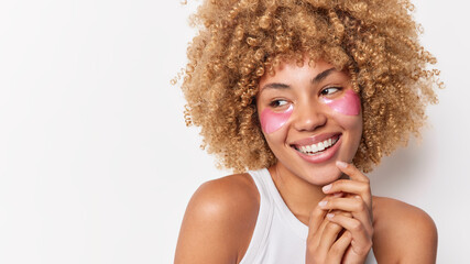 Horizontal shot of carefree cheerful woman concentrated away smiles toothily applies collagen patches under eye to remove fine lines stands bare shoulder isolated over white background copy space