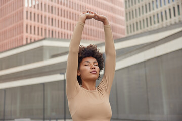 Relaxed woman breathes deeply meditates outdoors keeps arms raised up keeps eyes closed dressed in sportswear exercises in urban setting stretches outdoors. People wellness and physical activity
