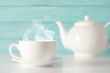 White cup with steam and teapot with herbal tea on blue wooden wall background