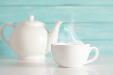 White cup with steam and teapot with herbal tea on blue wooden wall background