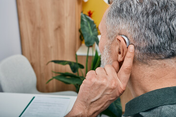 Hearing solutions for elderly deafness people. Older gray-haired man tunes his hearing aid behind the ear by pressing his finger on setting button while visit to hearing clinic - obrazy, fototapety, plakaty