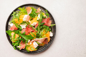 Summer melon salad with cantaloupe, prosciutto, soft cheese and arugula on white table background, top view, copy space