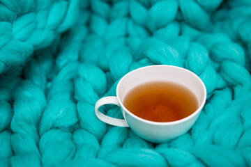 The concept of coziness and comfort is a green knitted blanket on which there is a white cup of hot tea. Place for an inscription.