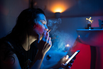 Young woman addicted to nicotine smoking cigarette and using mobile phone while sitting in bathroom under neon light, selective focus of depressed female reading about tobacco addiction on smartphone