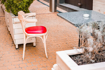 White and red classic soft chair stands near granite table with white ashtray. Traditional. Sidewalk cafe. Outdoor. Street. City. Restaurant. Sidewalk. Near building