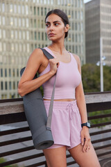 Vertical shot of thoughtful sporty woman dressed in tracksuit carries fitness mat concentrated away into distance poses outdoors against blurred urban background. Athletic female going to have pilates