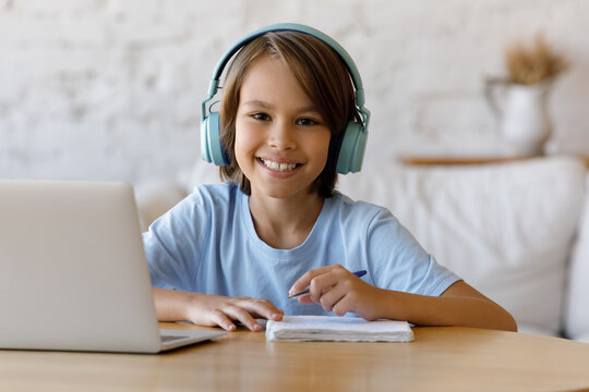 Happy school kid in trendy big headphones studying online from home, sitting at desk with laptop computer, writing learning notes, looking at camera, smiling, Homeschool, education on Internet - Powered by Adobe