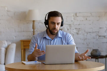 Serious male online teacher in headphones speaking at mic, using laptop, presenting tutorials to...