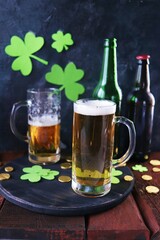 St. Patrick's Day, fresh beer in glass mugs and a bottle, gold coins, on a red wooden table, a green shamrock on a dark background, a party, a congratulation, a postcard