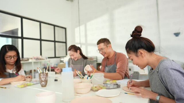 Group of Happy Asian man and woman friends enjoy and having fun indoor lifestyle painting self-made pottery together at pottery studio. Hobby leisure activity handmade ceramic art painting workshop.