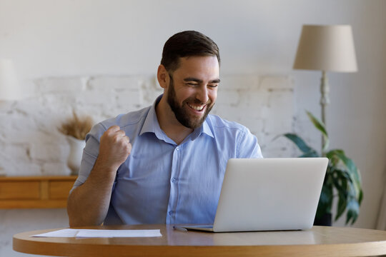 Excited happy millennial guy getting good news, looking at laptop display, smiling, reading email message, bank notice, laughing, making sinner gesture. Cheerful business man enjoying success