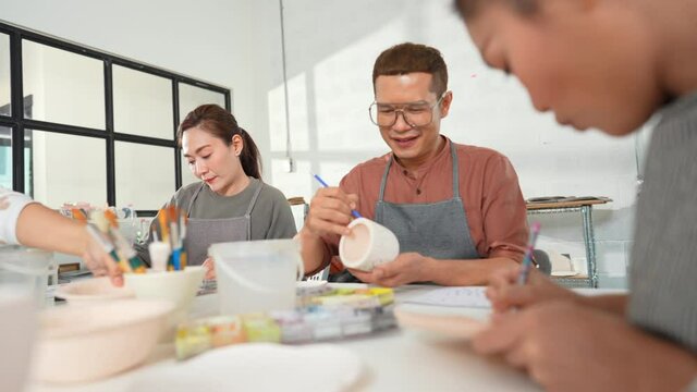 Group of Happy Asian man and woman friends enjoy and having fun indoor lifestyle painting self-made pottery together at pottery studio. Hobby leisure activity handmade ceramic art painting workshop.