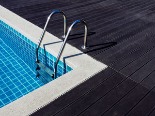 Stone edge frame on the square swimming pool with clean clear water, nobody. Square shaped pool with blue tiles with grab bars ladder on the dark wood decks, no people. Summer background.