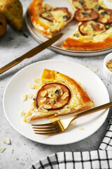 A slice of puff pastry pizza with pear, almonds, gorgonzola ricotta or blue cheese in a white plate on the kitchen table. Delicious layered pie with pear, dorblu, nuts on a light culinary background