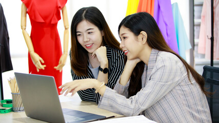 Two young businesswomen colleagues in working office style clothes looking at laptop computer screen together with happy action. Women in fashion and design business working on tailor shop