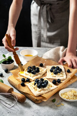 Obraz na płótnie Canvas Raw puff pastry with cheese cream and blueberries. The process of making delicious sweet buns or mini pies. Greasing the edges of the baking dough with egg. Hands in the frame 