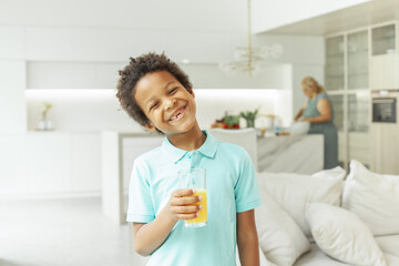 Cute little boy holding glass of orange juice and drinking at home