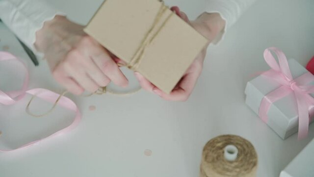Woman designer hands wearing thin shirt wrap cardboard box with present using thin cord tied in bow on table surface closeup