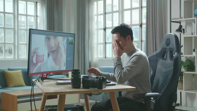 Asian Cameraman In Long Sleeved T-Shirt And Black Pants Being Tired While Using Desktop Computer For Editing Photo At Home.
