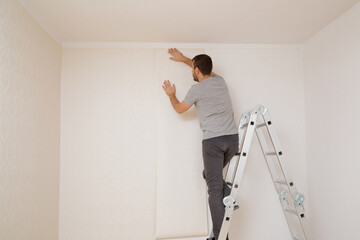 Young adult man standing on metal ladder and applying new wallpaper on white wall in room. Repair...