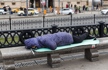 homeless man sleeps on a bench in the city center against background of the train station. Moscow, Russia