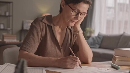 Waist up shot of mid-adult Caucasian woman at desk taking notes in study materials, studying from home in evening
