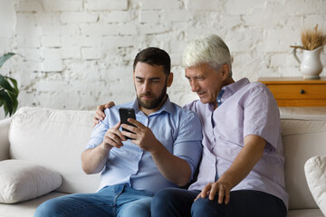 Focused grownup son teaching mature father to use online virtual app on smartphone, internet...