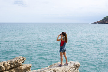Girl with jean shorts and sneakers standing in a rock photographing the mediterranean sea during her vacations. Summer leisure and hobbies.