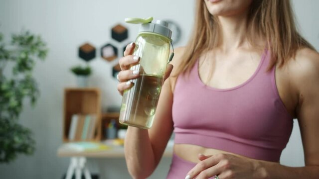 Tilt-up portrait of attractive girl opening bottle and drinking water then looking at camera and smiling indoors in apartment. People and healthy lifestyle concept.