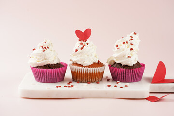 Three homemade white and chocolate muffins with white creamy frosting and heart-shared red and white sugar decoration on marble board on pink surface