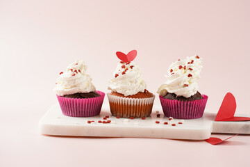 Three homemade white and chocolate muffins with white creamy frosting and heart-shared red and white sugar decoration on marble board on pink surface