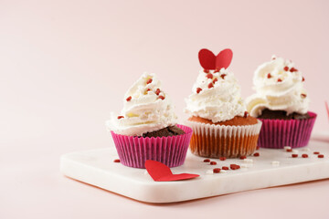 Three homemade white and chocolate muffins with white creamy frosting and heart-shared red and white sugar decoration on marble board on pink surface