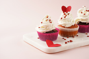 Three homemade white and chocolate muffins with white creamy frosting and heart-shared red and white sugar decoration on marble board on pink surface