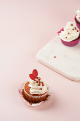 Three homemade white and chocolate muffins with white creamy frosting and heart-shared red and white sugar decoration on marble board on pink surface