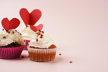 Three homemade white and chocolate muffins with white creamy frosting and heart-shared red and white sugar decoration on marble board on pink surface