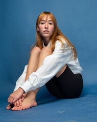 Fototapeta na wymiar Sitting on the floor girl with red hair and white skin isolated on blue in white shirt and black breeches