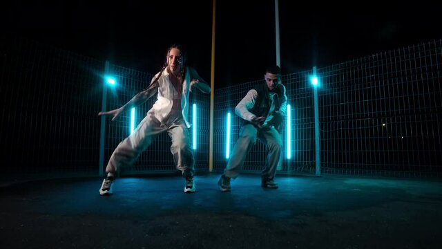 Duet of dancers enjoying hip hop moves at night city with neon light at background. Stylish group of two dancers performing afrohouse. Urban view. Street dance. Afrodance. Slow motion in 4K, UHD