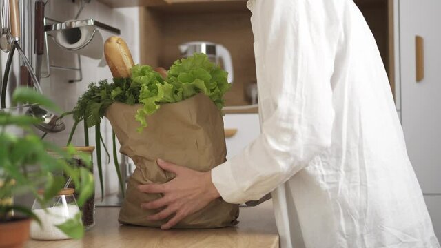 A young hostess brings a bag full of healthy products. Modern home delivery. Various vegetables and bread in eco paper bags that a young girl put on the kitchen table at home. Eco supermarket
