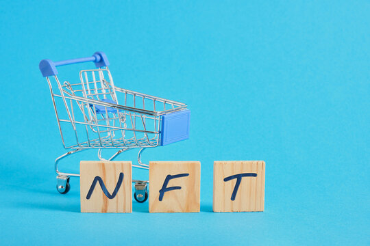 wooden blocks with the letters nft and a miniature shopping cart on a blue background - Powered by Adobe