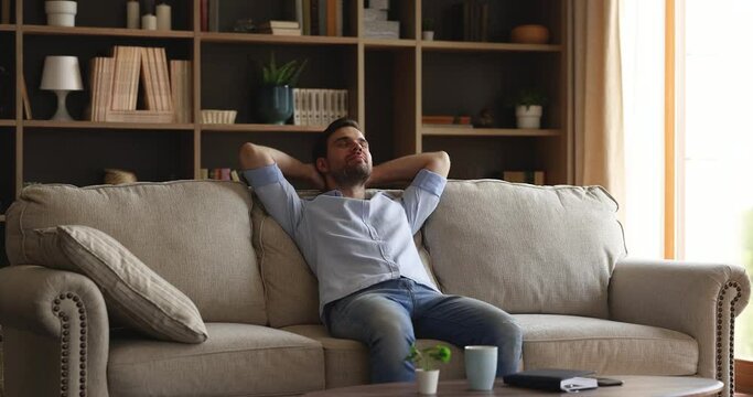 No stress, happy owner of fashionable, comfort apartment concept. Young serene man relaxing on cozy sofa inside modern living room, put hands behind head resting on couch, breath fresh conditioner air