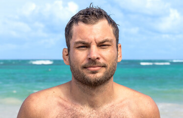 Handsome individual shirtless adult man white ethnicity portrait, confident man standing on the beach and looking at the camera, the sea in the background.