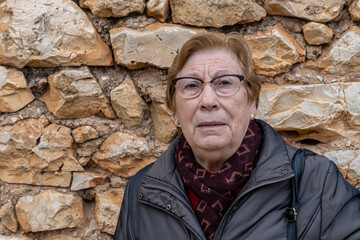 Close-up portrait of an older Caucasian woman wearing glasses and wearing a trench coat, with a stone wall in the background.