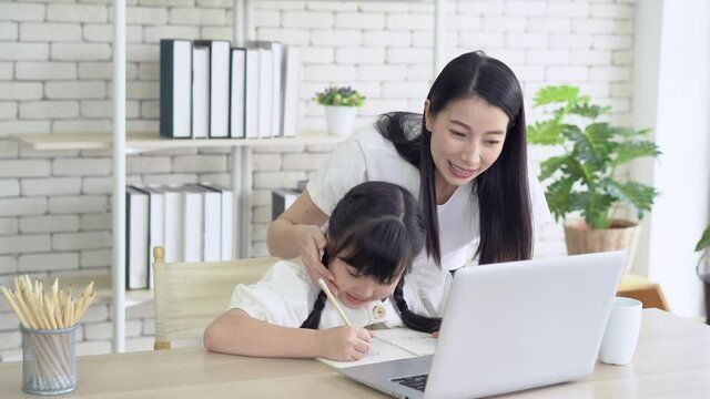 Portrait of young Asian mother helping and teach her daughter with homework while sitting on desk at home. Mom braids hair daughter and smile. Little girl happy to study at home education.