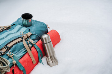A tourist backpack lies on the snow, a red mat is fastened on the side, a thermos is next to it....