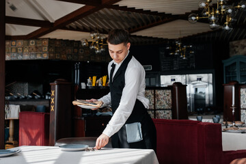 A young male waiter in a stylish uniform is engaged in serving the table in a beautiful gourmet restaurant. A high-level restaurant. Table service in the restaurant.
