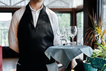 A young waiter stands with wine glasses in a stylish modern restaurant. Table service in the restaurant. Haute cuisine, service and maintenance. Close-up.