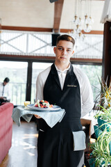 A young waiter in a stylish uniform carries an exquisite salad to a client in a beautiful gourmet...